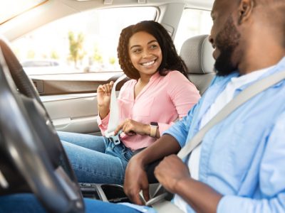 Safety First. Smiling African American Couple Fastening Seatbelts Before Driving In City, Sitting Inside New Luxury Car, Following Safe Ride Rules, Ready For Travel With Auto, Looking At Each Other